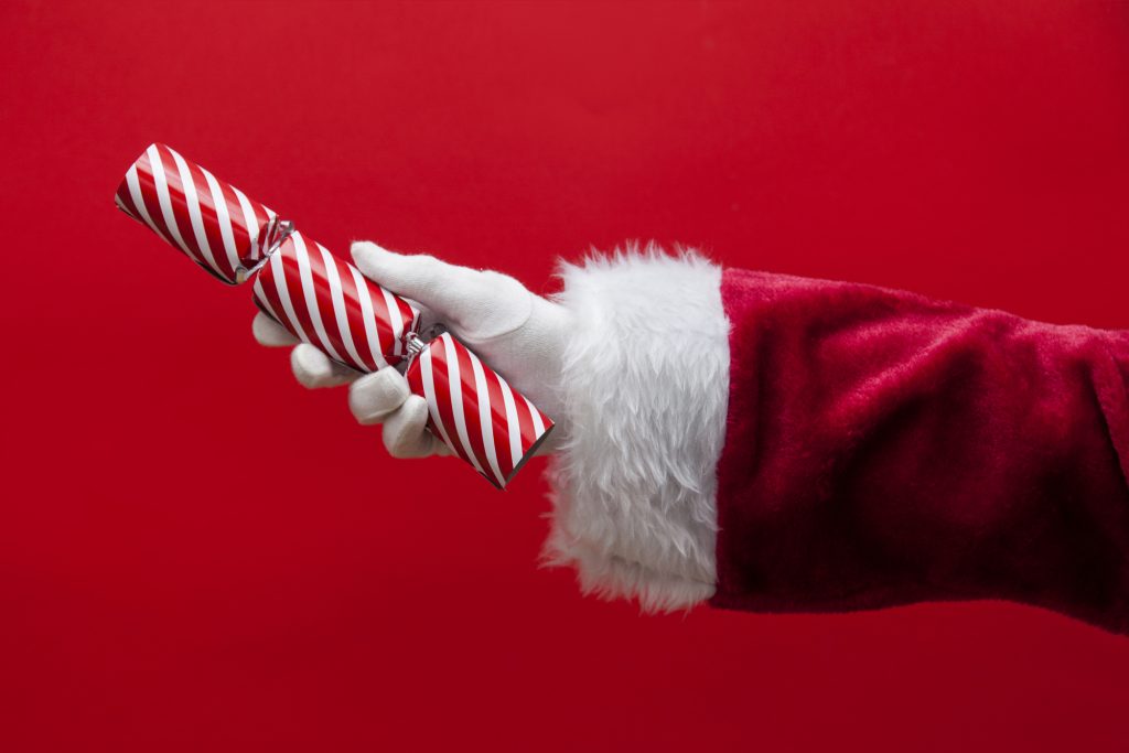 Un Père Noël tenant dans sa main un Christmas cracker. Ajoutez une touche anglaise à votre Noël avec les Christmas crackers. Surprenez vos proches avec cette tradition festive pleine de surprises