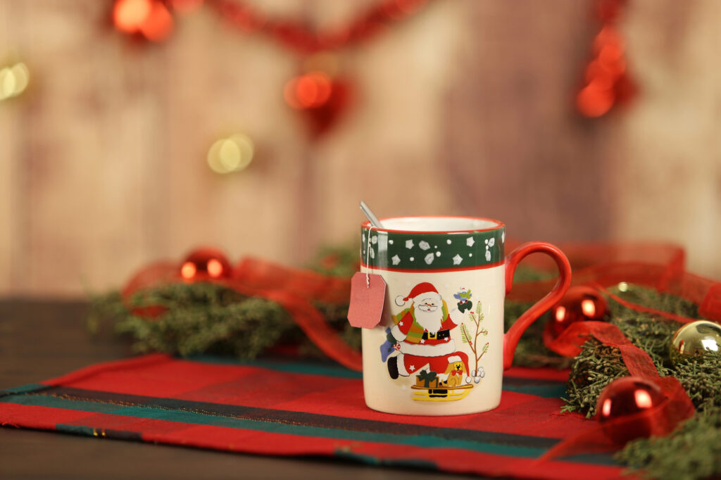 Mug de Noël décoré d’un Père Noël, posé sur une nappe rouge parmi des boules et des branches de sapin