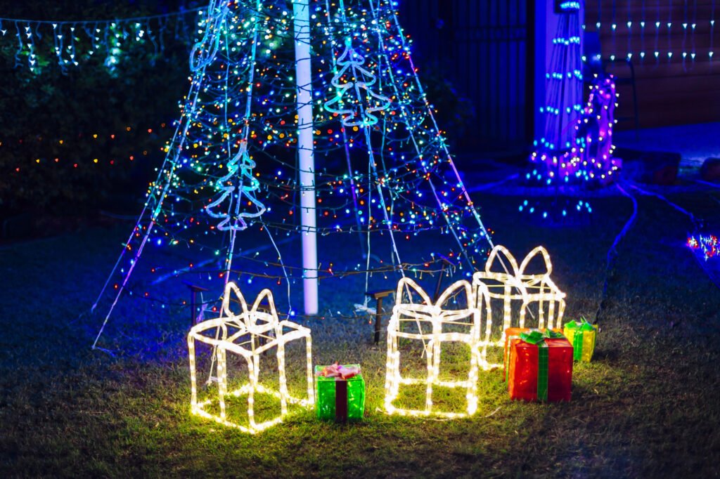 Jardin décoré pour Noël la nuit : sapin lumineux, boîtes à cadeaux éclairées et guirlandes sur la façade