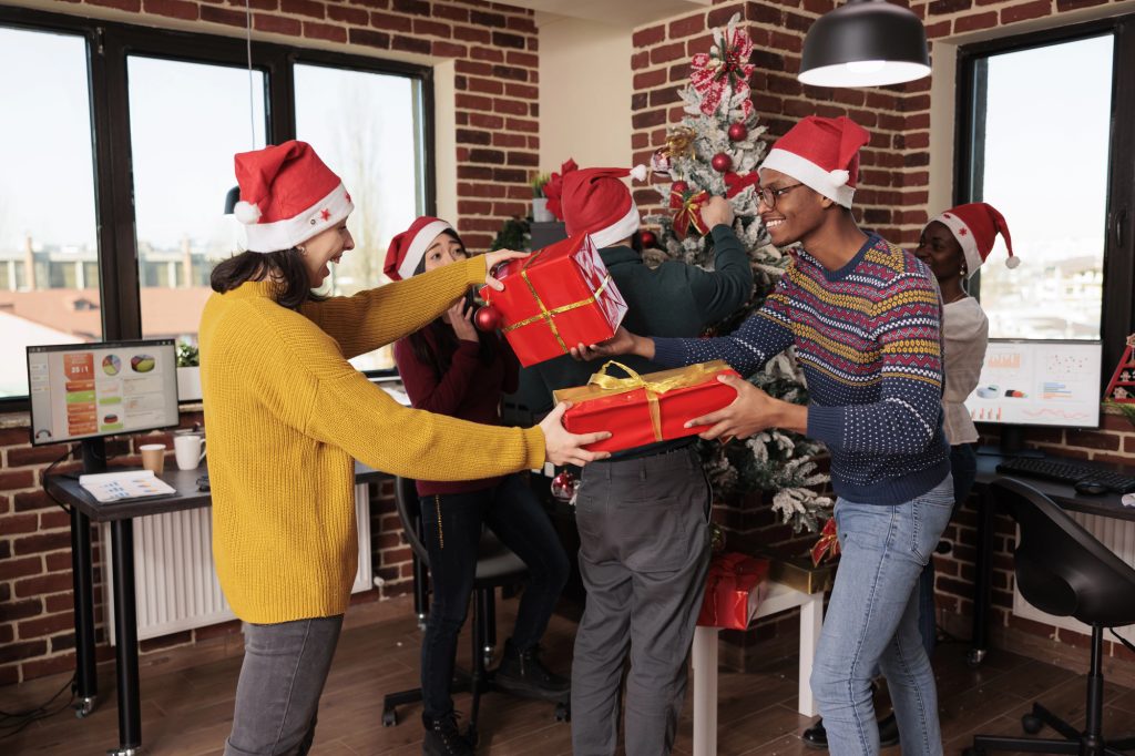 Collègues en bonnet de Père Noël s'échangeant des cadeaux emballés de rouge devant le sapin du bureau