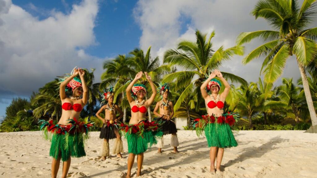Danseuses polynésiennes en costume traditionnel et coiffe de fleurs sur une plage de sable blanc bordée de cocotiers