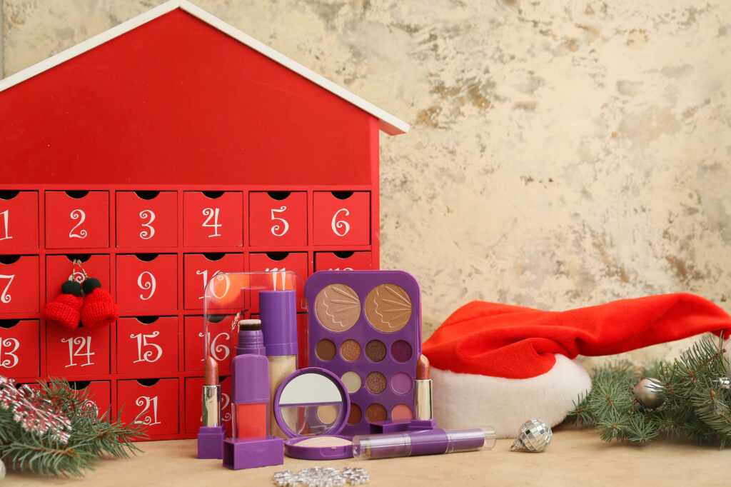 Calendrier de l'Avent beauté en bois rouge à tiroirs numérotés, entouré de maquillage violet et d'un bonnet de Père Noël